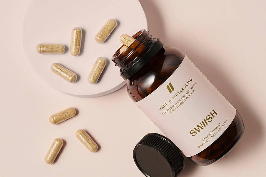 Bottle of SWIISH Hair + Metabolism supplement with lid open and capsules spilling out on a beige background
