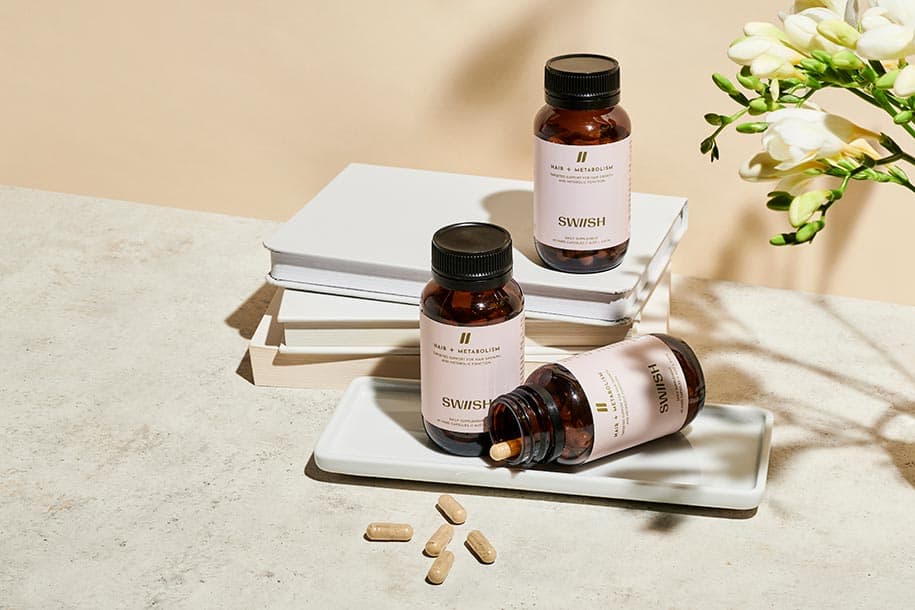Three bottles of SWIISH Hair + Metabolism supplements styled with a stack of books and fresh flowers
